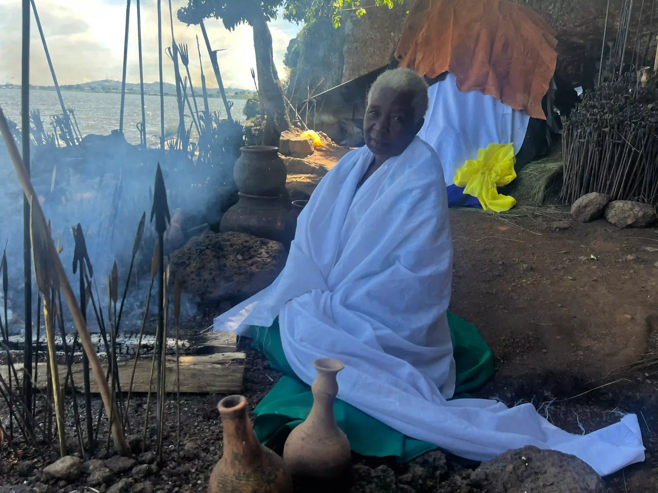 Mama Tezira in her waterside spiritual consultation space with traditional ceremonial pottery, incense, and ritual healing items